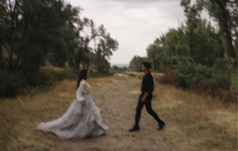 A slightly blurry photo of an engaged couple walking towards each other. They are outdoors with tall greenery framing them.