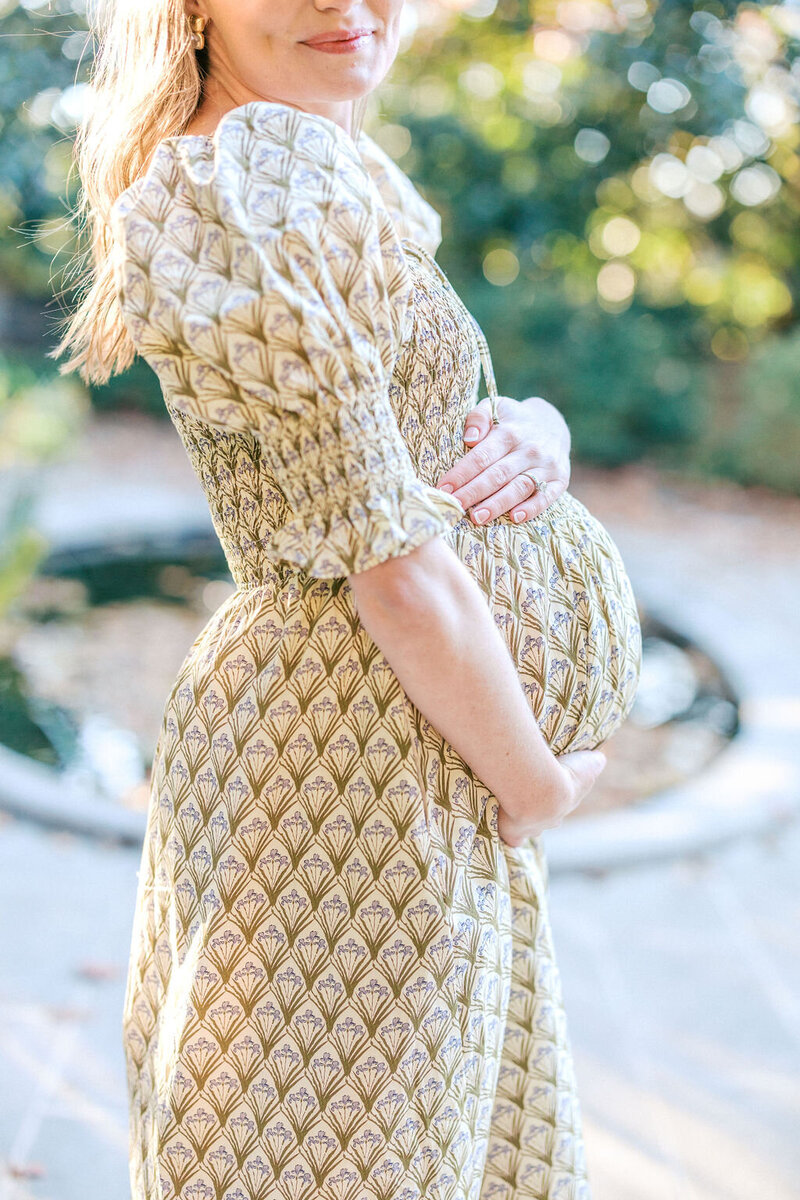 Woman in green patterned dress holds baby bump with gardens behind her
