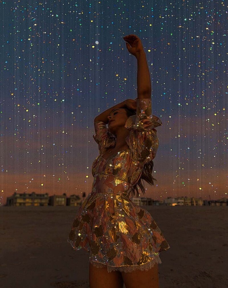 Under a starry night sky, a woman in a shimmering dress stands with her arm raised and eyes closed, creating a dreamy, magical scene. This image is for Debra LeClair's Embodied Style services.