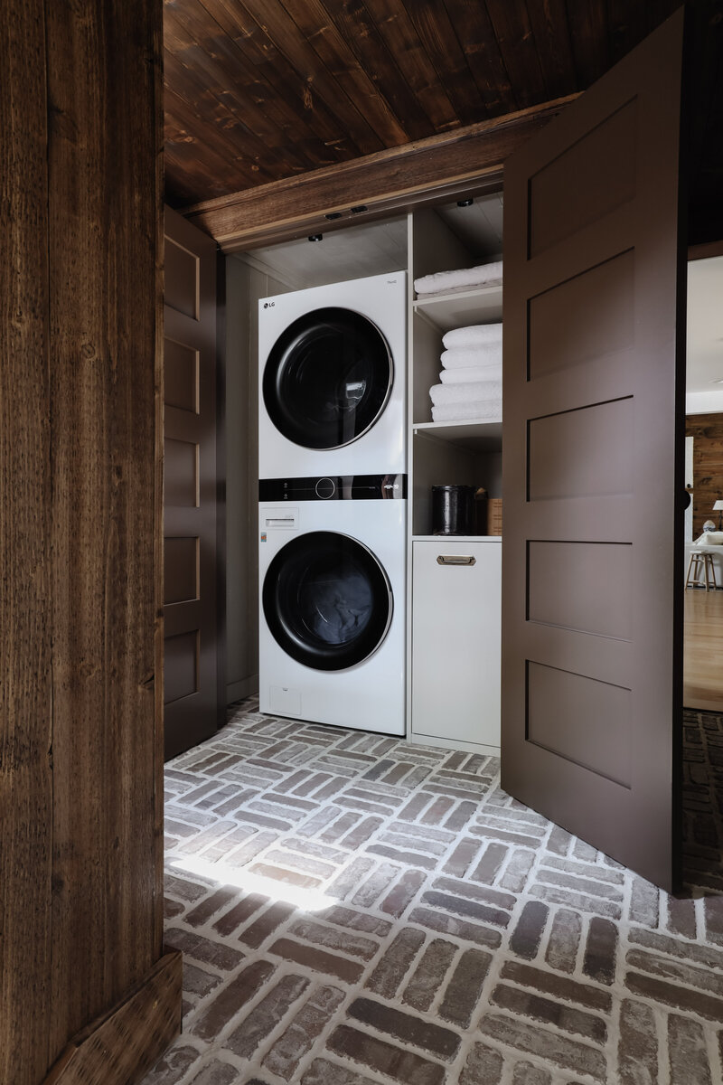 Moody Rustic Laundry Room renovation by Nadine Stay | Dark wood stained walls. Brown painted doors.  DIY inset laundry hamper drawer. Basketweave brick floors. Washtower and laundry closet organization.