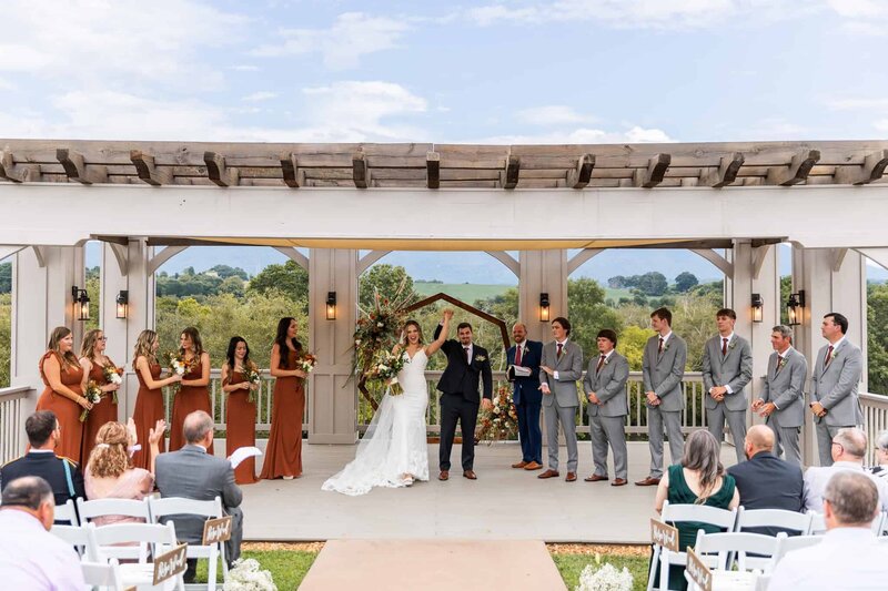 On a mountain-facing ceremony platform, a bride and groom raise their joined hands triumphantly just after saying “I do.” Rust-colored bridesmaid dresses and lush florals frame them, with the hazy blue Appalachian ridge in the distance. It’s a stunning, natural setting that mirrors many beloved ceremony sites in Pennsylvania’s own mountain regions.
