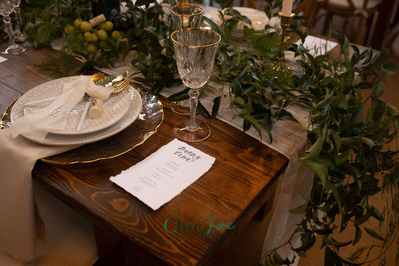 Place setting featuring a menu on a rectangular wooden farmhouse table with gold-rimmed glassware, ceramic plates, and greenery accents.