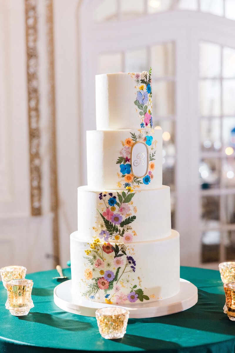 A multi-tiered wedding cake with colorful floral designs and monogram initials on a table, surrounded by glass votive candle holders, meticulously arranged by an Iowa wedding planner.