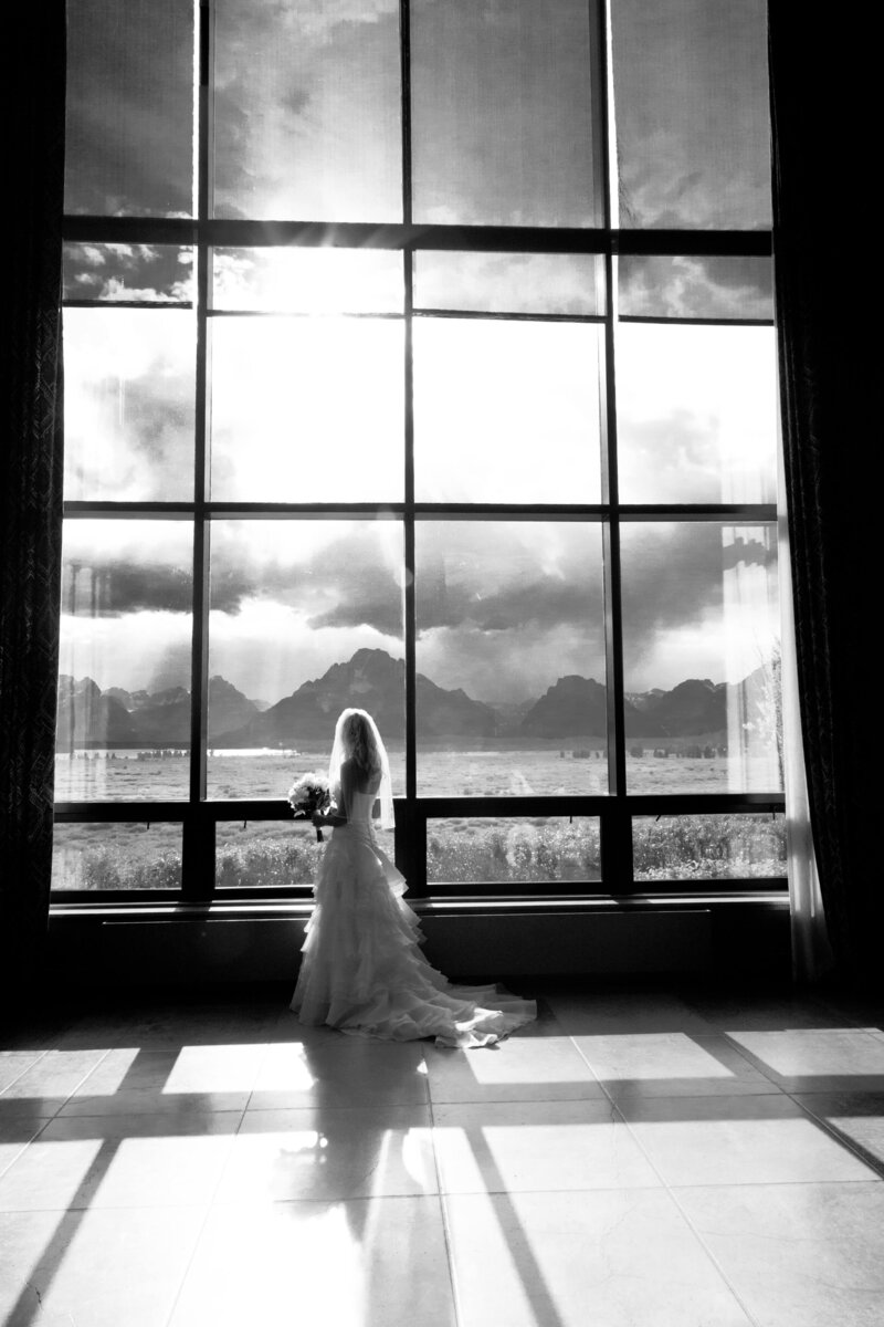 Black and white image of a bride standing in front of the floor-to-ceiling windows at Jackson Lake Lodge, gazing at the Grand Teton National Park landscape as sunlight streams through.