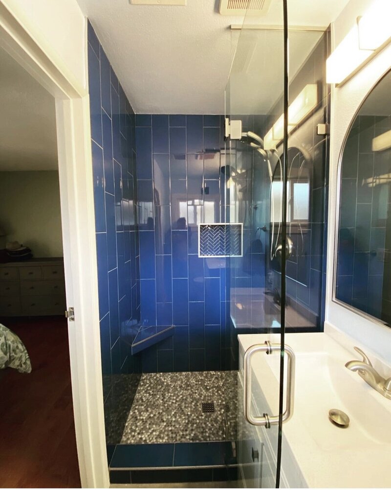 A remodeled shower with blue vertical tiles and white and grey stones as the floor of the shower. There is a vanity with white countertops and brushed nickel hardware.
