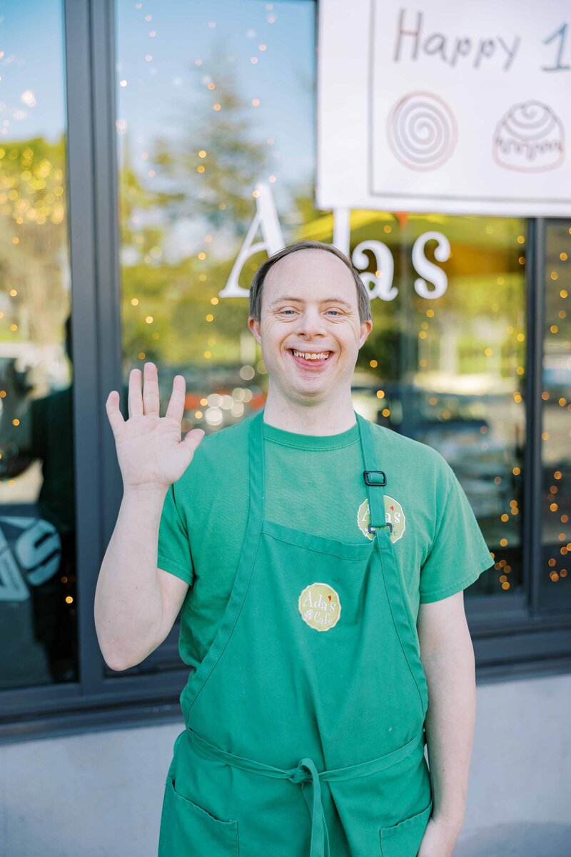 Man wearing a green apron that says "Ada's Cafe" on it and smiling at the camera
