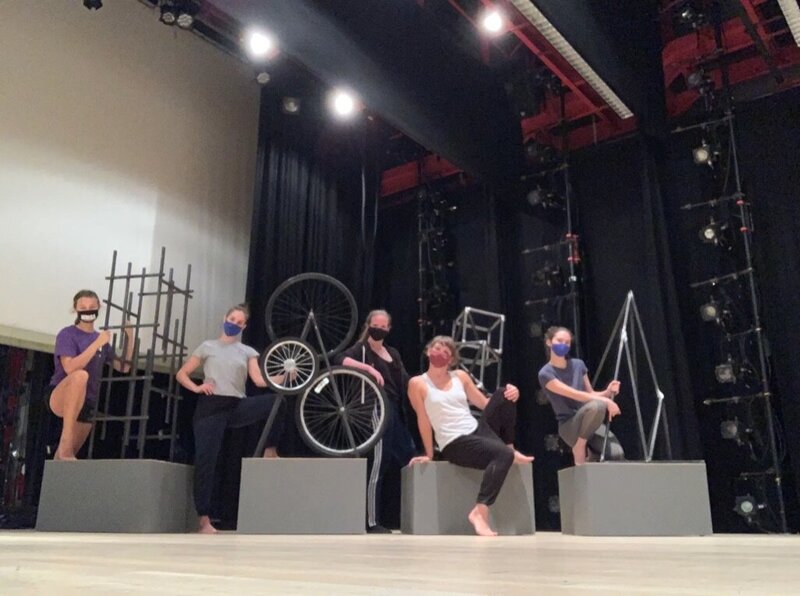 Dancers wearing masks and facing the camera. Sitting on prop blocks or leaning up against prop bicycle wheels