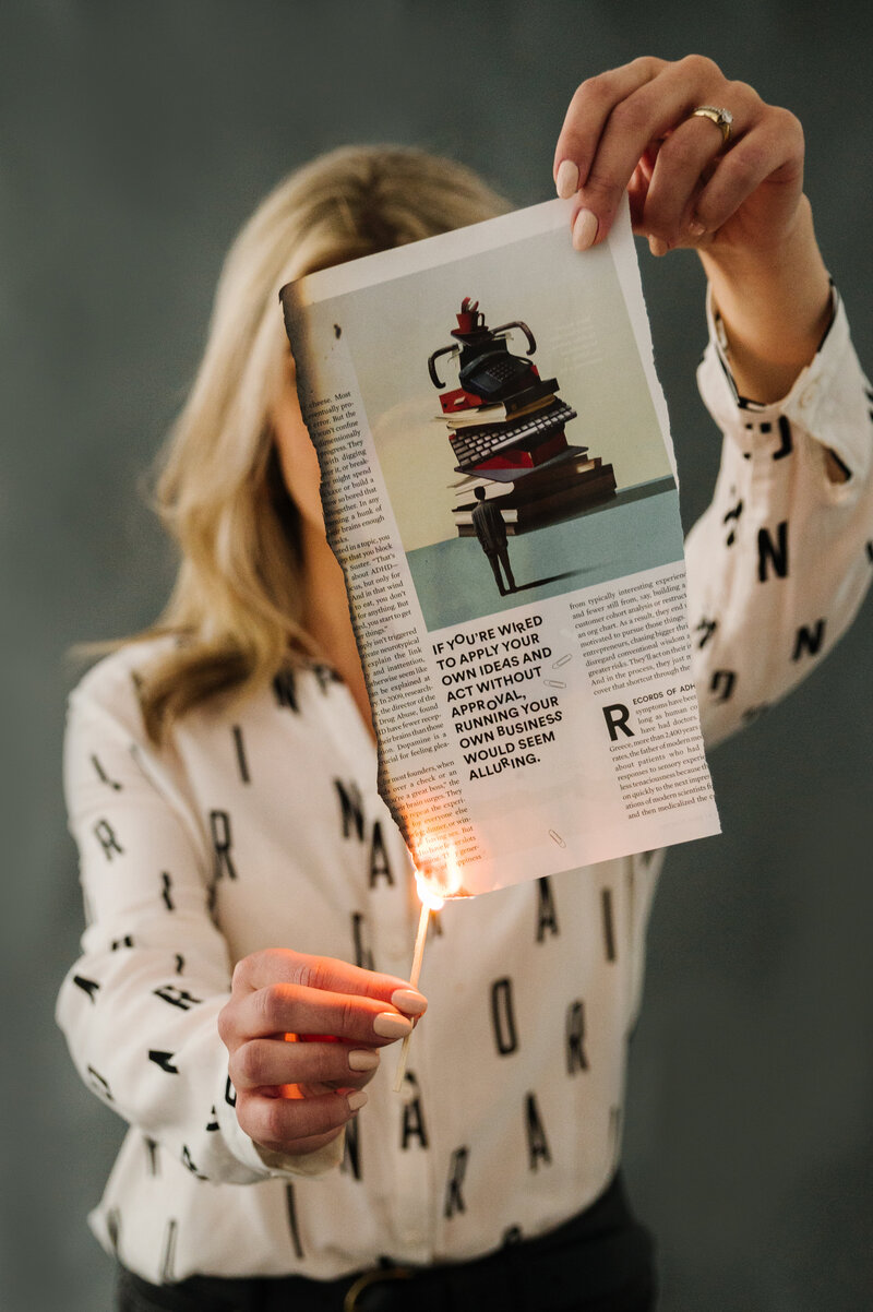 Sarah Klongerbo sitting on a stool in front of a black background with magazine pages hanging with clothespins on string behind her