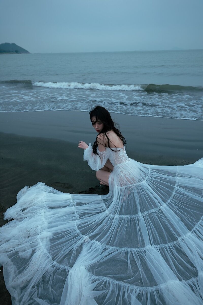 A woman wearing a delicate sheer couture dress shot on the beach in dark moody tones.