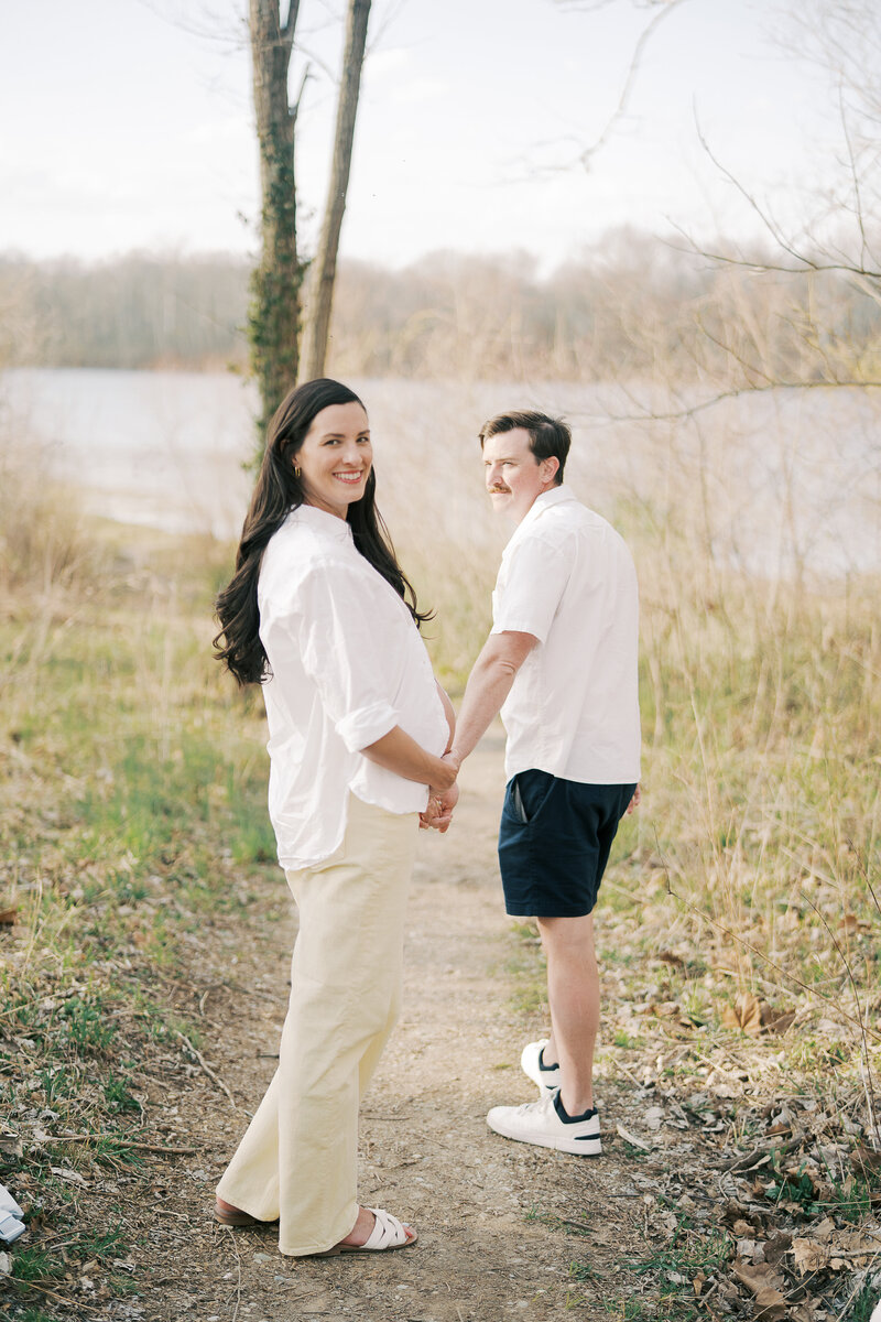 a pregnant woman holding her husband's hand while they walk near a lake