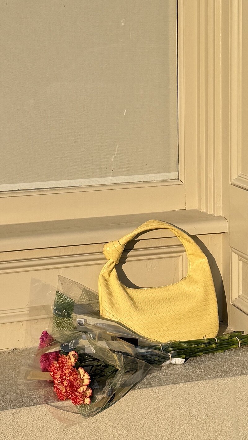 A yellow handbag sits on a sunlit windowsill next to a bouquet of red and pink flowers wrapped in plastic. The scene feels warm and serene.