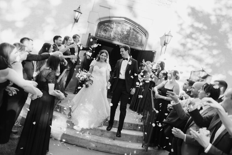 black and white image of bride and groom walking down the steps after ceremony while guests throw rose petals image by wedding photographers in minnesota tom thornton photography