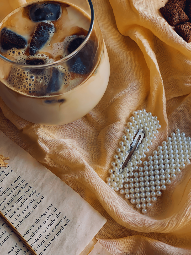 A glass of iced coffee with visible coffee cubes, pearl hair clips, an open book, and a soft yellow linen fabric, creating a cozy and elegant aesthetic.