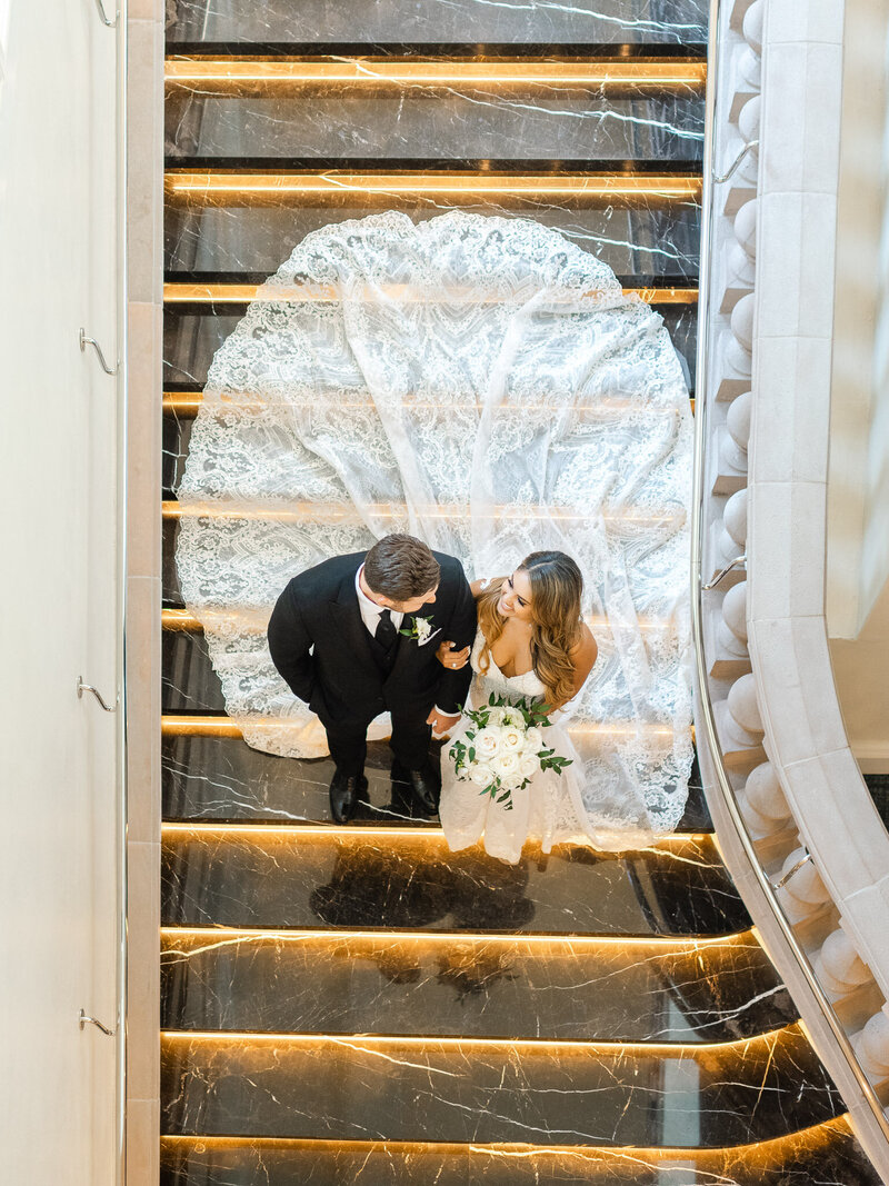 A newlywed couple standing on the grand staircase at Ritz-Carlton Laguna Niguel, with the bride’s lace train fanned out behind her in an elegant display