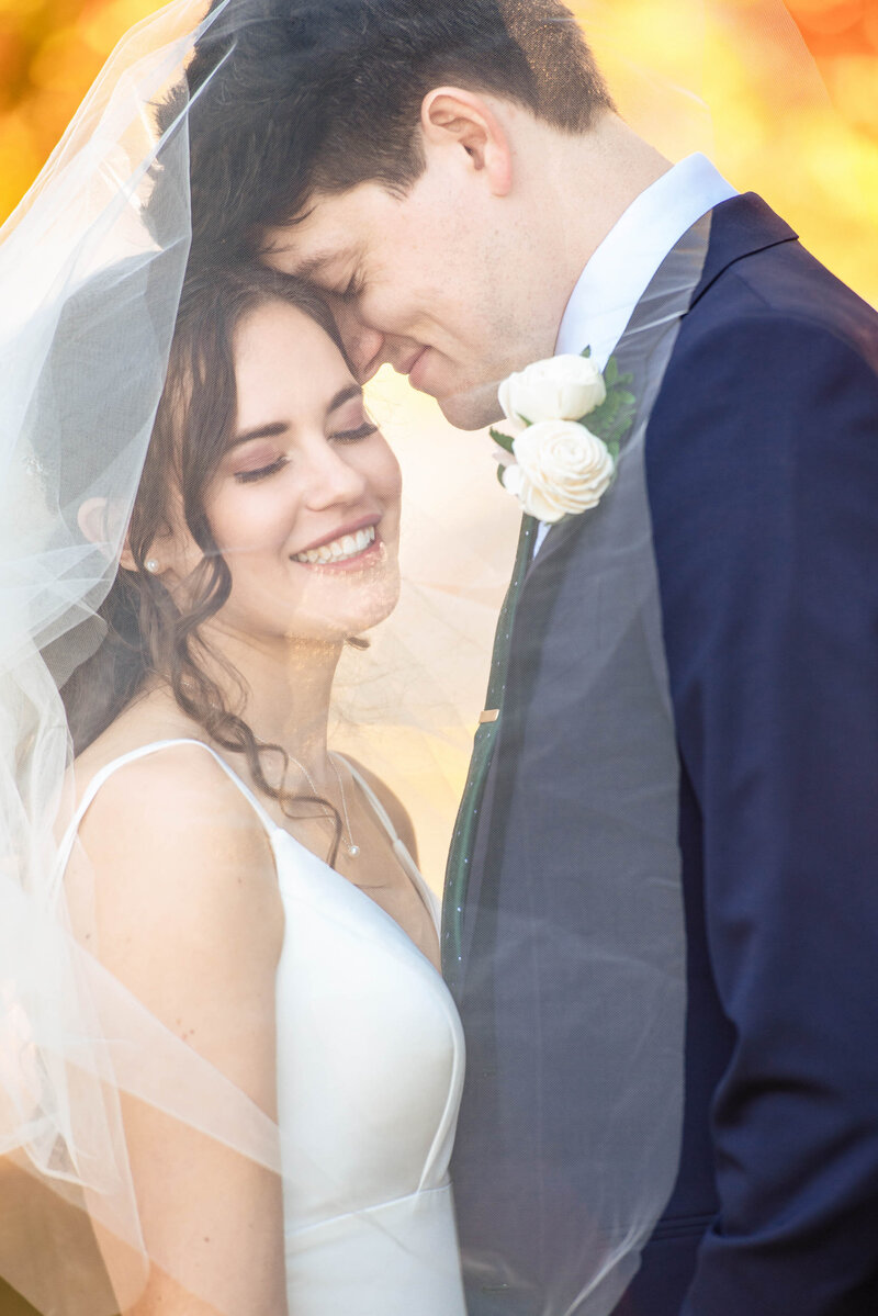 Bride and groom snuggled underneath the brides veil with their eyes closed and smiling with bright yellow autumn leaves blurred in the background  at Freedom Park by Charlotte wedding photographers DeLong Photography