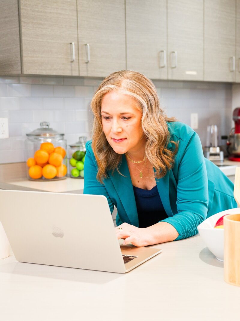 Woman in teal blazer leaning on the kitchen counter working on her laptop.