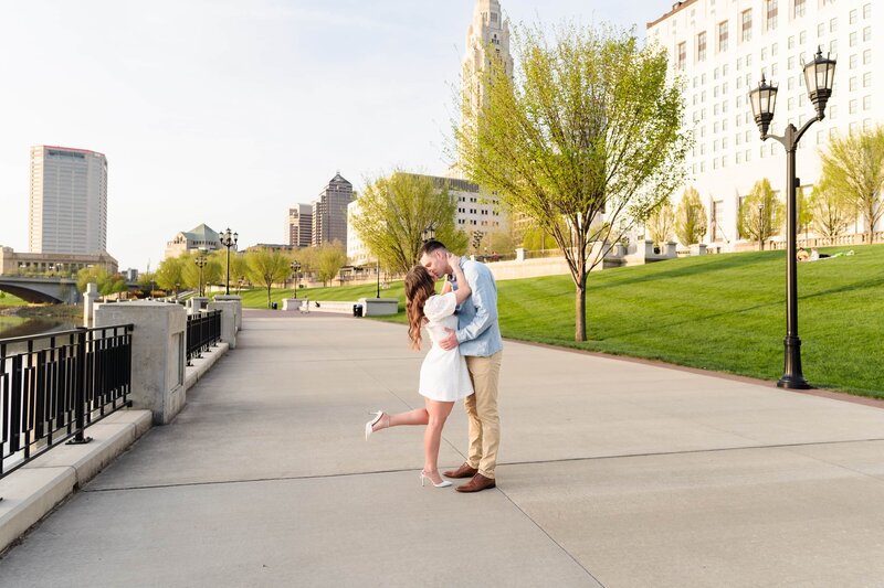 A young man in a light blue shirt and a young woman in a white dress kiss along the Scioto Mile with the Columbus skyline in the background