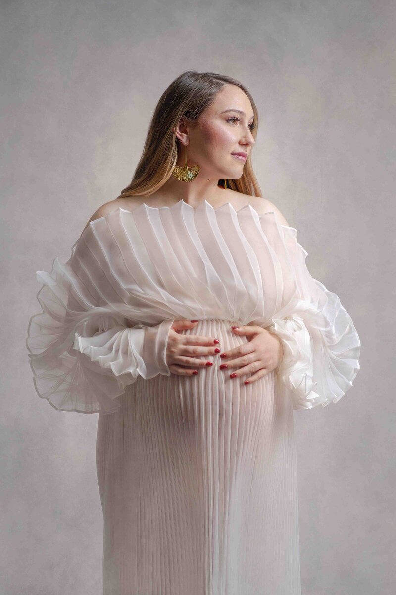 Bellevue Mom poses in pregnancy photo session for maternity photographer Lana Stewart in a white pleated dress that is semi-transparent. The sleeves are almost ruffled due to the pleating. She has a gold gingko earring and red nail polish.