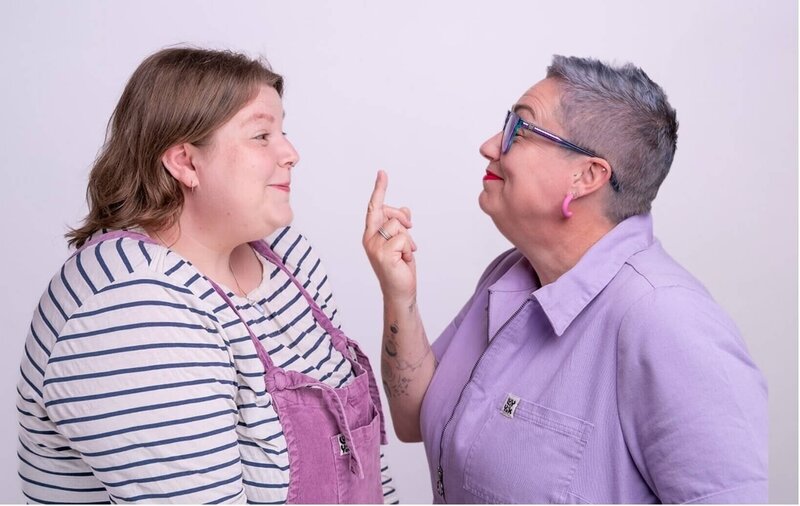 Pippa on the right, in a lilac jumpsuit, smiles and playfully holds a middle finger up at Lucy, who’s wearing a striped top and purple dungarees, during a Disobedient Business® Co. photoshoot.