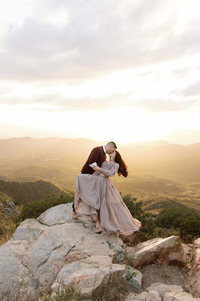 Man dips and kisses woman on the edge of a cliff at sunset