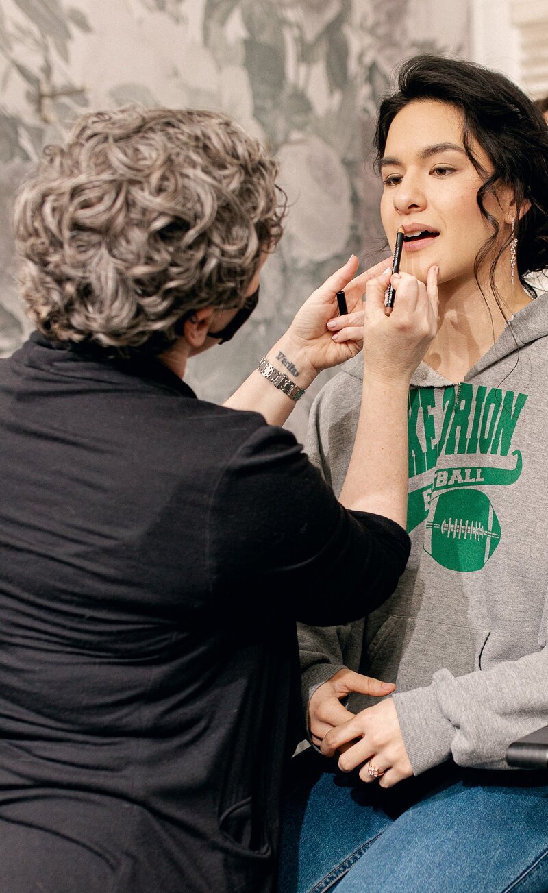 The makeup artist applies lip liner to a model with dark hair, full brows and delicate skin.