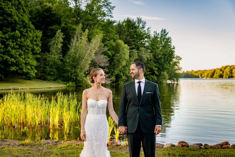 A couple holding hands and looking at each other while standing in front of a lake.
