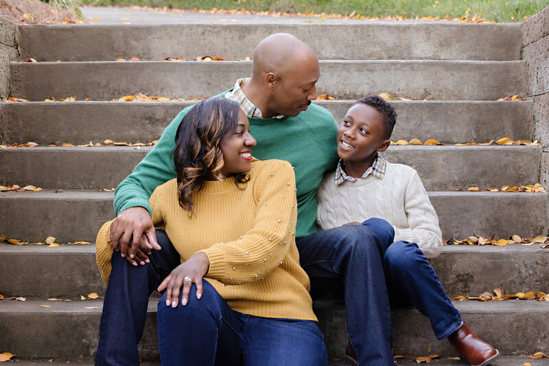 Sweet family sitting on concrete steps with golden autumn leaves scattered around. Mom has a yellow sweater, Dad has a greenish teal sweater and the son has an oatmeal colored sweater. All wearing jeans for casual, stylish family photos.