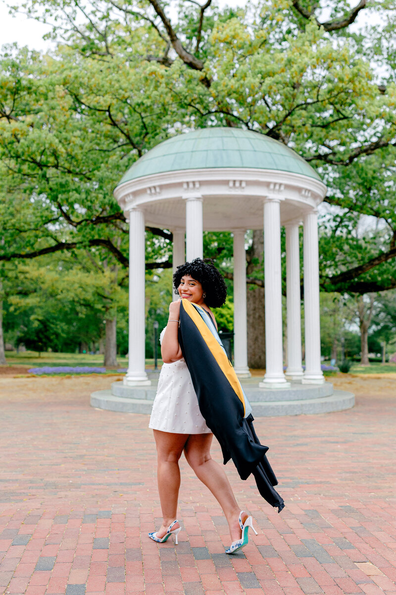 UNC graduate in white dress smiling at the camera while walking past the Old Well with cap and hood over shoulder.