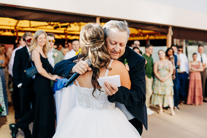 A bride in a white dress hugs an older man in a suit, who holds a microphone and a piece of paper, during an outdoor wedding celebration. Guests stand in the background, watching the emotional moment captured by an Austin photographer.

