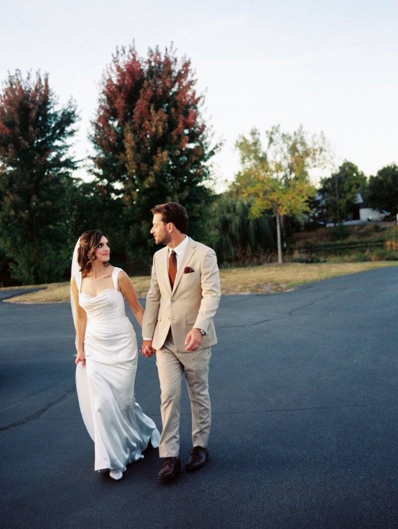 image by tom thornton photography of bride and groom dressed for minnesota wedding walk along road holding hands looking at each other with trees in background