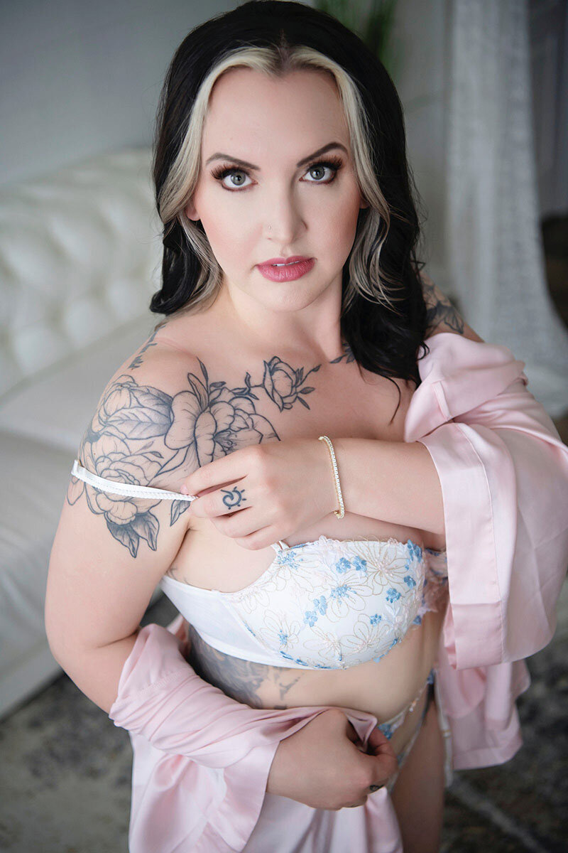 Woman in floral lingerie and pink robe, standing confidently with a serene expression. Tattoos on shoulder, styled hair; elegant, intimate setting.