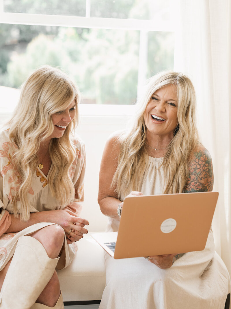two women sitting in window seat looking at  laptop