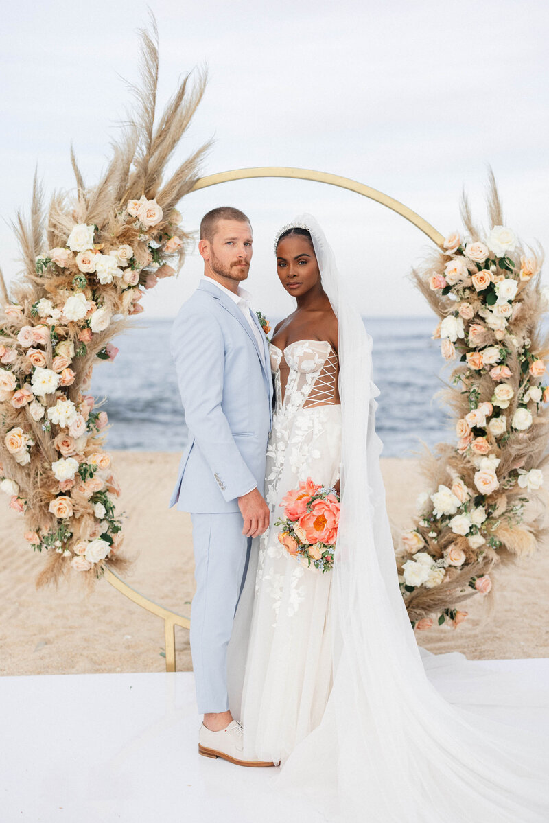 A white groom and a black bride pose in front of a circular structure decorated with white and peach flowers and dried pampas
