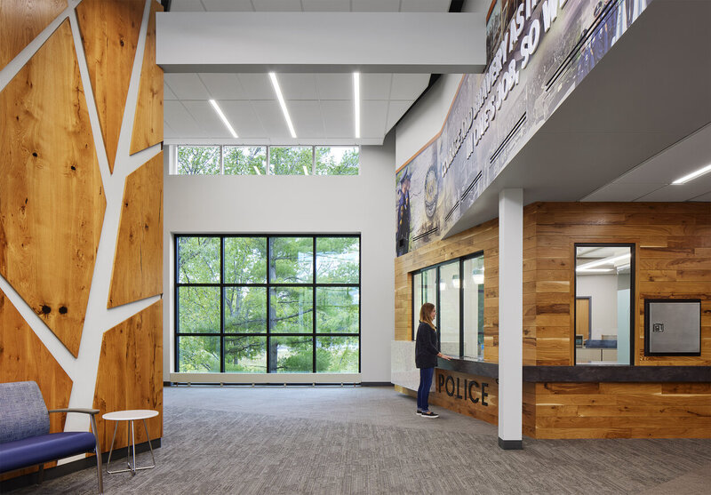 Lobby of a police station with large windows letting in natural light, wood and glass service counters, and a person standing at the reception area marked 'POLICE'.