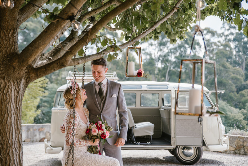 Bride and groom with bouquet sitting on swing with white kombi wedding car in background