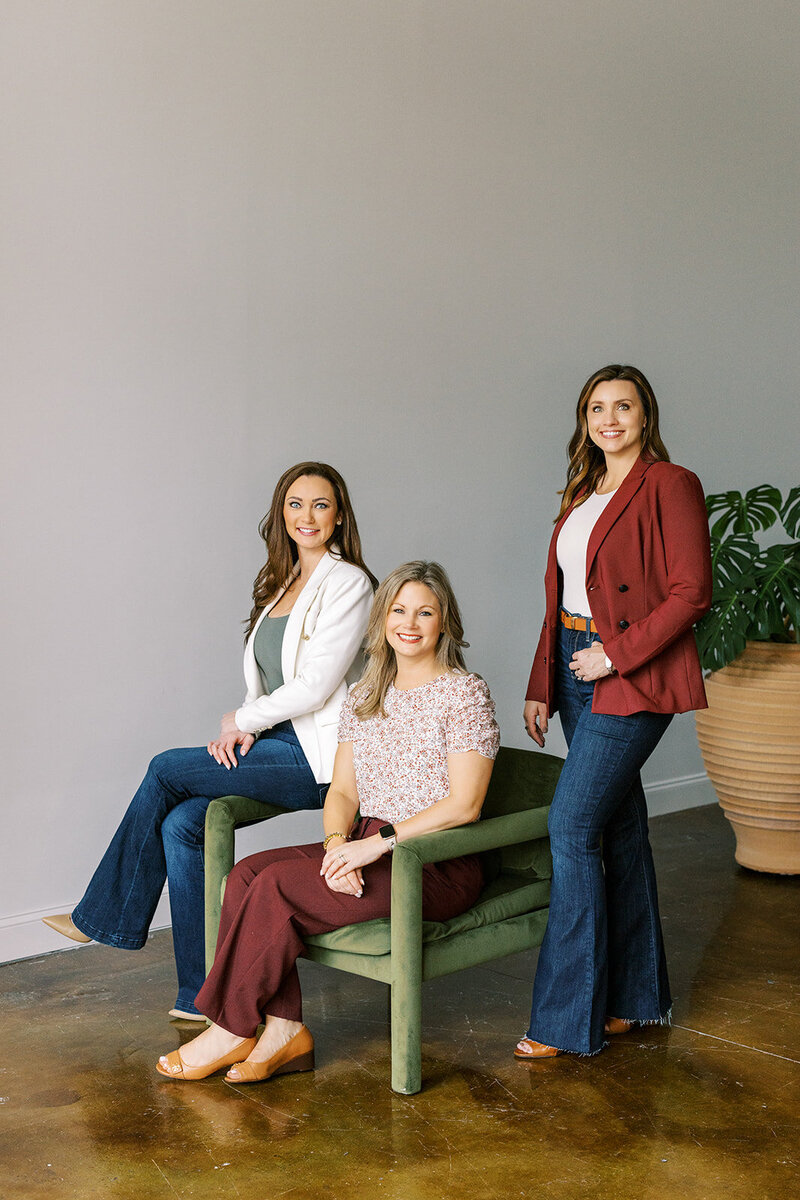 Three women are posing in a light-filled room. One is seated in a green chair wearing a pink top and dark pants, while the other two stand beside her in blazers and jeans. A large potted plant is in the background. They all smile at the camera.