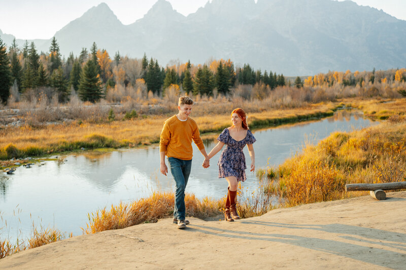 Grand Teton National Park photographer, Adriana Montoya Photo. A couple, holding hands and walking towards the camera with the Tetons in the far distance behind them. The girl is looking at the guy while he is looking ahead at the ground. Both are smiling. Right behind them is a river and trees.