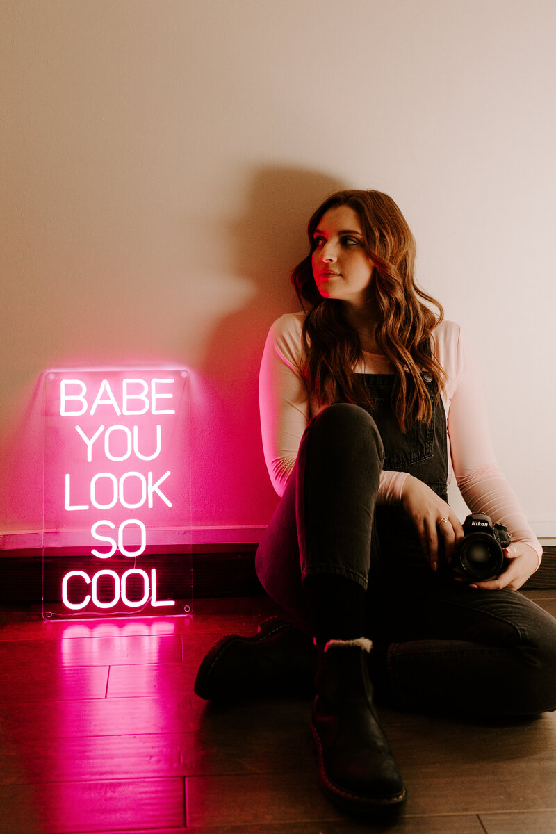 A portrait of the wedding and couples photographer sitting next to a pink neon sign that reads " Babe You Look So Cool". She is also holding her digital camera and looking away from it.