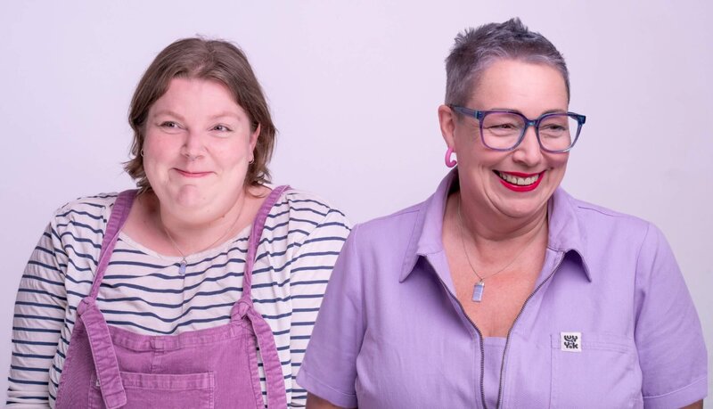 Two people stand side by side against a pale lilac backdrop. Lucy on the left wears a striped white and navy top with mauve dungarees and smirks; Pippa on the right has cropped purple-grey hair, blue rimmed glasses, pink hoop earrings, red lipstick, and a lilac collared shirt, laughing.