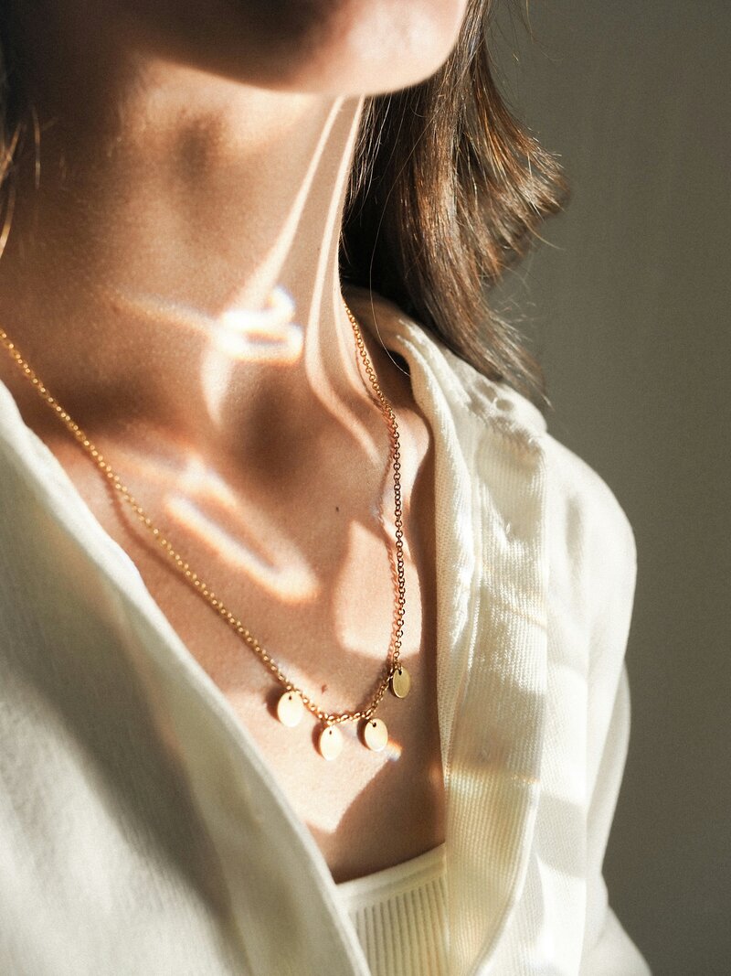 Two-layer gold charm necklaces on a woman’s neck with soft shadows and sunlight.