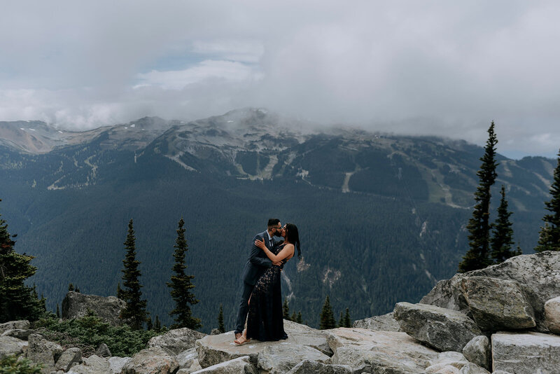 A couple standing on a dock with mountains in the background kiss and hold each other