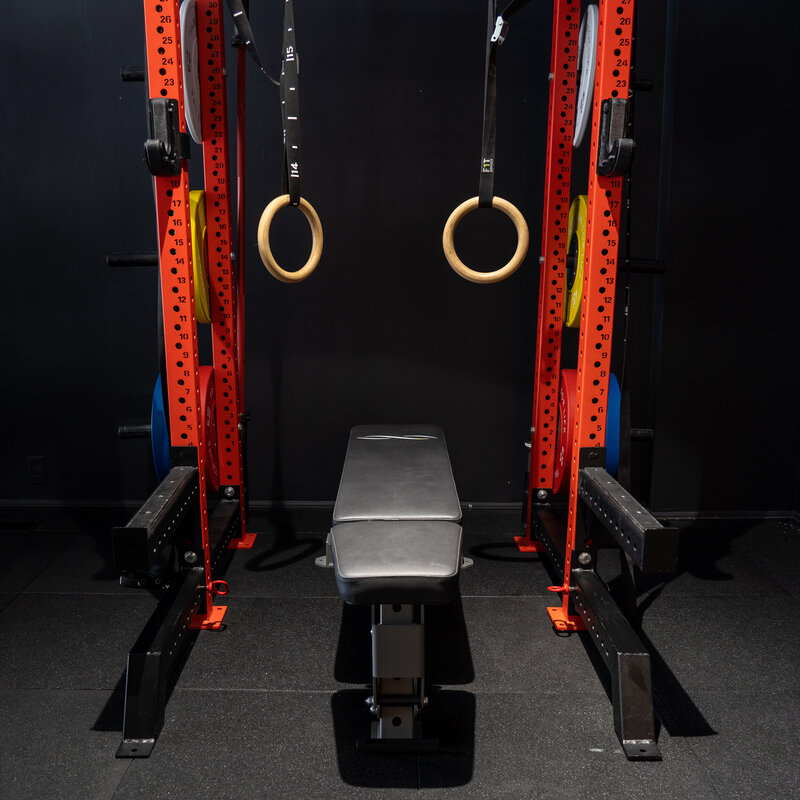 A photo of one of the red squat racks at Train For Life's Personal Training Gym in Collingwood Ontario. The red squat rack has a pair of gymastics rings hanging off the rack.  There is a bench sitting between the rack.