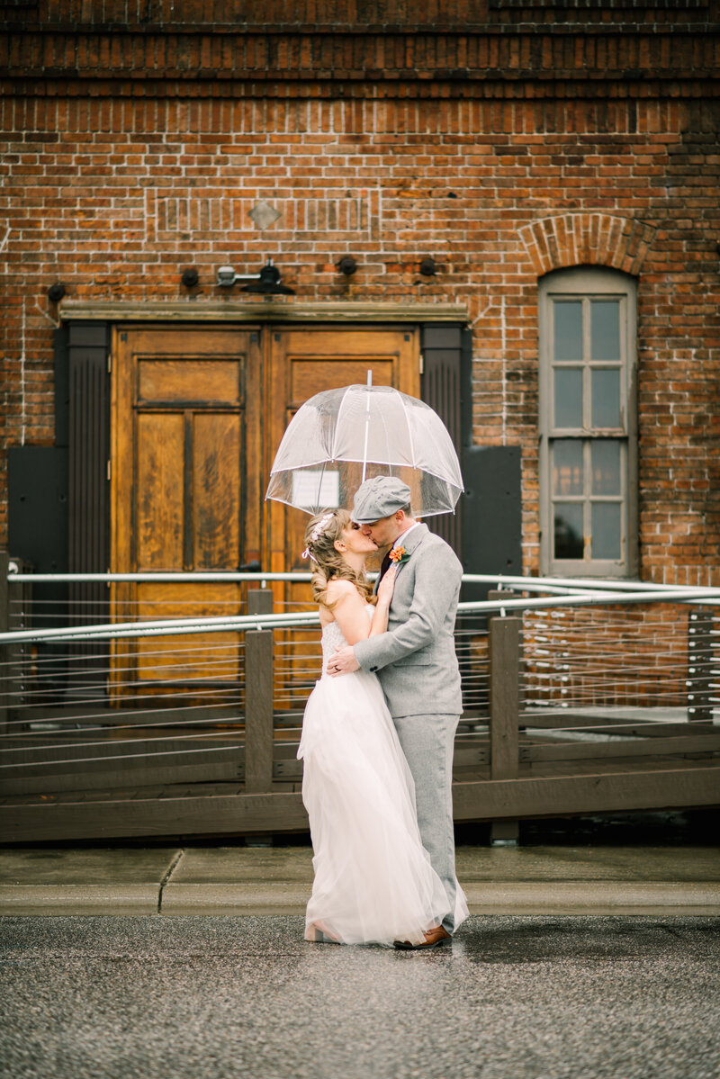 Bride and groom kiss under an umbrella in the rain.