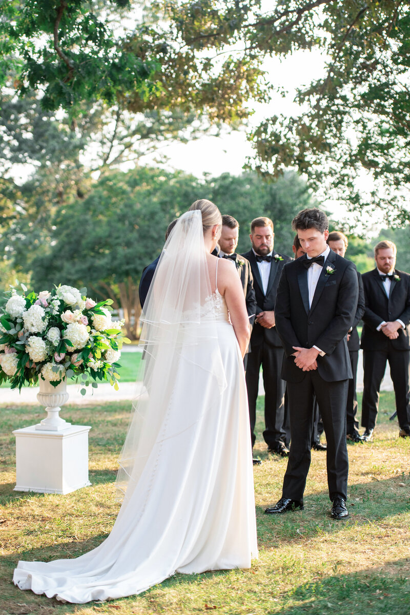 A bride in a white gown and veil stands beside a groom in a black suit during an outdoor wedding ceremony. They are surrounded by groomsmen in formal attire and a large floral arrangement. The setting is a grassy area with trees in the background.