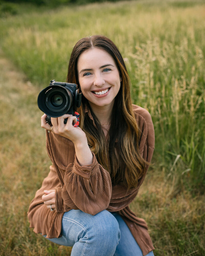 Photographer, Kim Pickett, sits in a field in nature while holding a camera and smiling