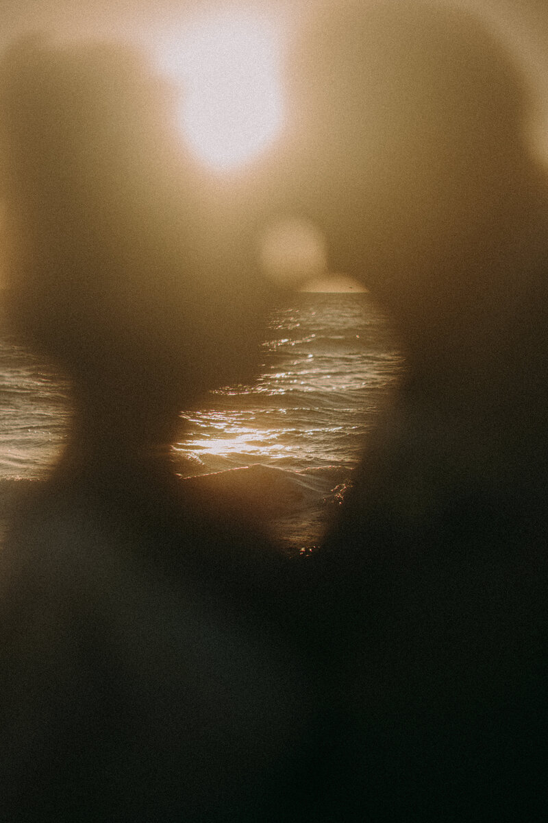 A bride and groom rest their heads on each other. The camera blurs them out and focuses on the ocean behind them.
