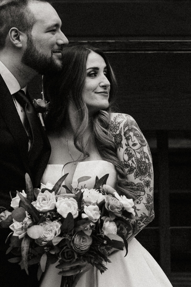 A black-and-white image of a couple standing close together, one person adorned with a floral tattoo and holding a bouquet of roses in Strawberry Hills Arizona.