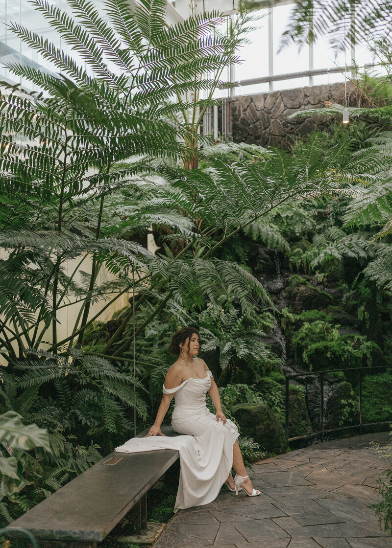 A bride sits on a bench underneath tropical palms.