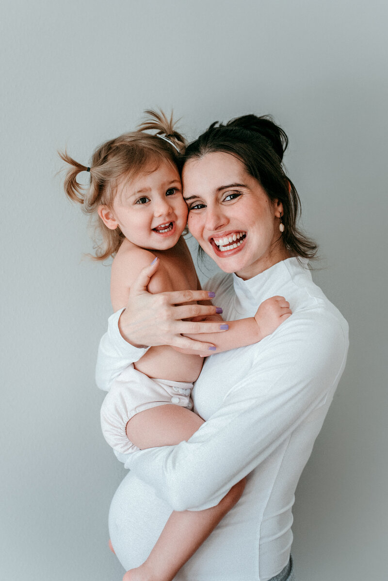 pregnant mother wearing white high neck and long sleeve bodysuit holding her toddler girl with pony tails, both with huge smiles looking at the camera