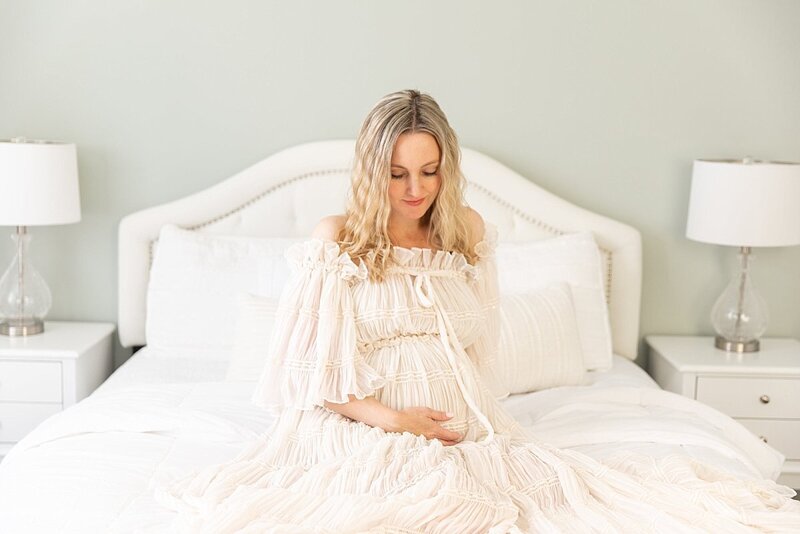 Pregnant woman in cream off the shoulder dress on bed by Maryland Maternity Photographer : Rebecca Leigh Photography